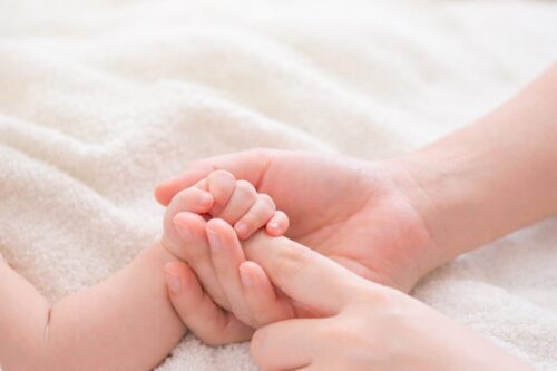 赤ちゃんの小さな手を大人の手でやさしく包み込んでいる様子。白いタオルの上で行うベビーマッサージのイメージ写真。