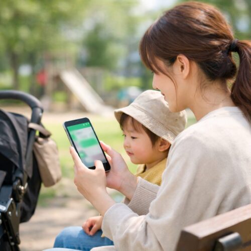 公園のベンチで子どもと一緒にスマートフォンを見るママの後ろ姿。画面はぼかされ、子連れのお出かけ中にLINEを使うイメージ写真